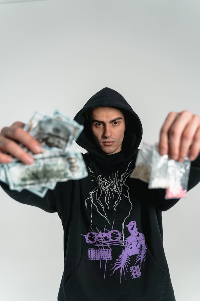 Young man in hoodie holding money and bags with a serious expression, illustrating illicit trade.